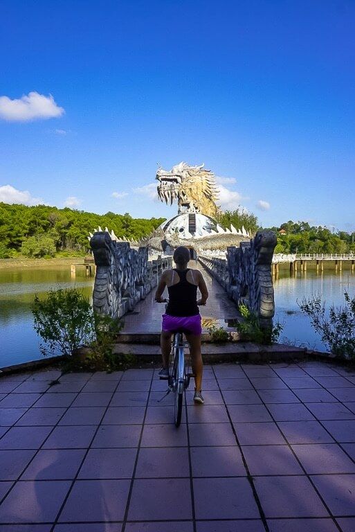 Kristen on bicycle in front of narrow bridge leading to abandoned dragon building hue water park