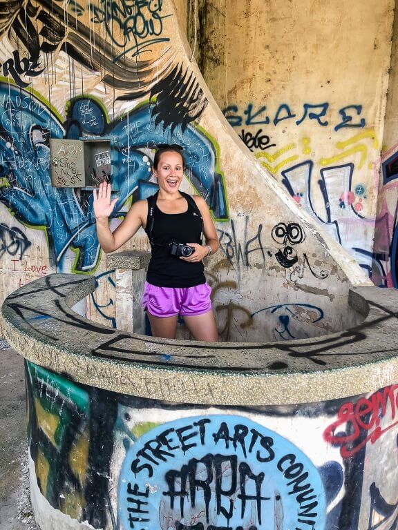 Kristen stood behind a circular stone bar waving surrounded by graffiti