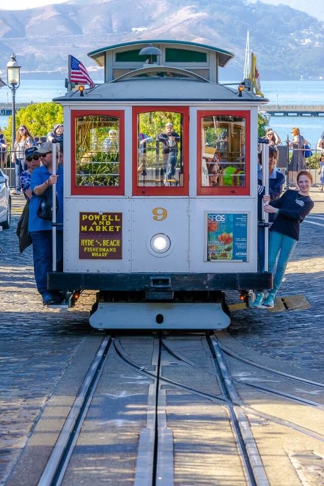 famous cable car of san francisco with tourists hanging off the sides san francisco bay in background