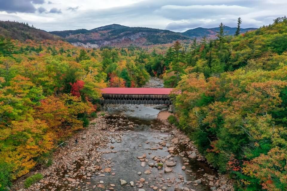Drone picture of covered bridge crossing river on kancamagus highway in new hampshire america stunning red bridge surrounded by trees and hills in background