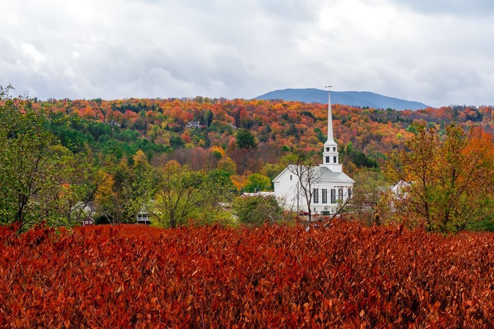 Famous white church in stowe vermont with stunning burnt red foliage in foreground and hills in background filled with trees amazing pictures of america