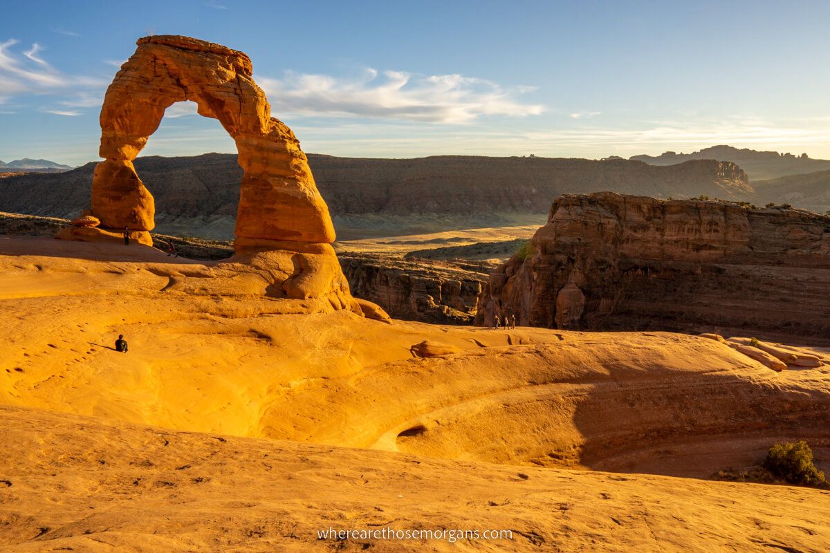 Photo of a wide open red rock landscape with a giant natural arch next to a deep bowl-shaped depression with sun rays bursting in from the side in Arches National Park