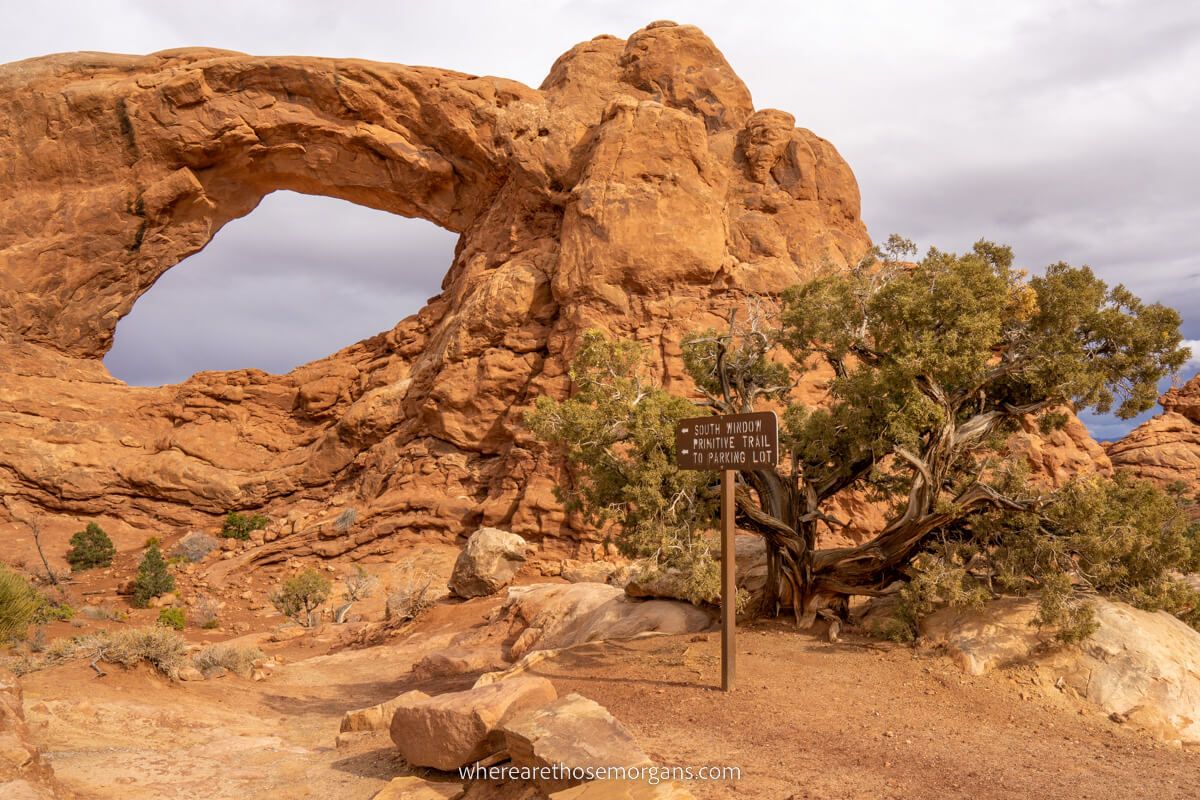 Photo of a window shaped hole in a massive red rock formation with a tree in the foreground next to a trail sign