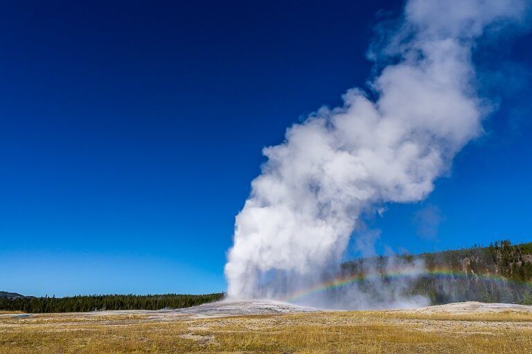 Old Faithful geyser erupting white steam against a deep blue sky and rainbow appearing