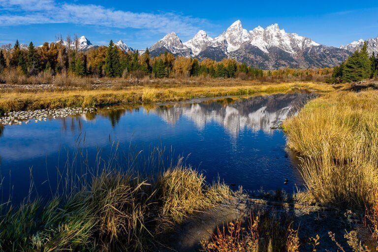 Snow capped mountains reflecting in almost still river with grass and trees surrounding