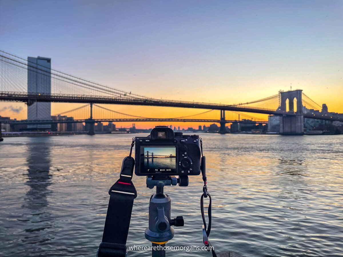Camera on a tripod taking a photo of the Brooklyn Bridge and East River from Pier 17 in NYC at sunrise