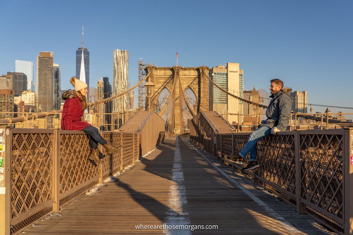 Mark and Kristen Morgan from Where Are Those Morgans sat on low metal fences on either side of the Brooklyn Bridge at sunrise with Manhattan's skyline in the background