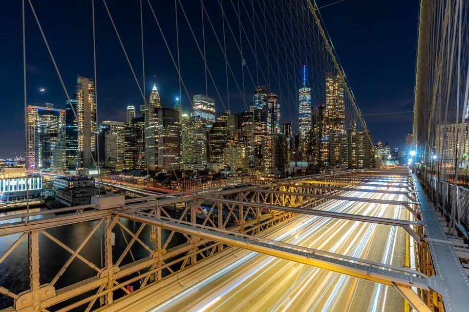 Cars driving across a bridge featuring a long exposure blur effect with lights and manhattan skyline in background at night