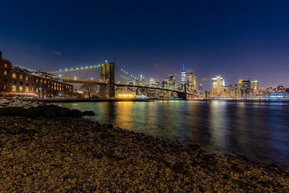 Brooklyn Bridge at night photography taken from Pebble Beach in Brooklyn colors reflecting in river in NYC