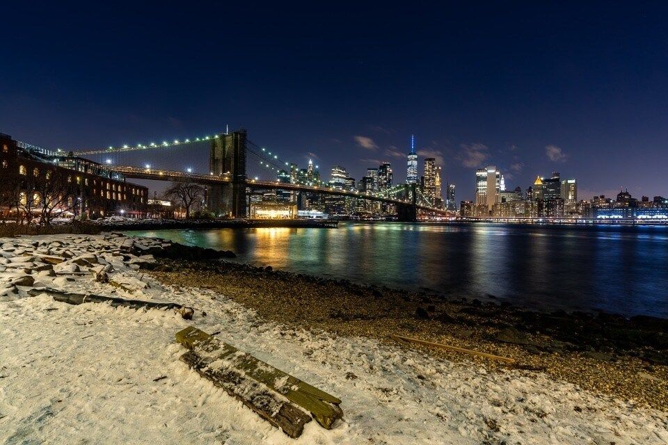 Pebble Beach view of janes carousel time out market and nyc skyline in background behind Brooklyn bridge with snow on beach