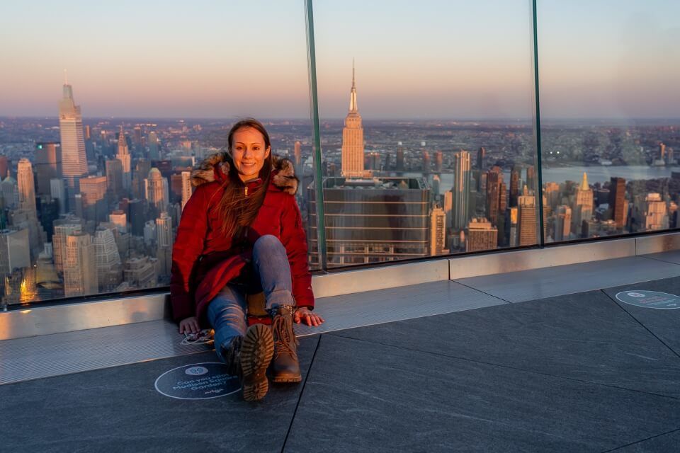 Woman sitting on the Edge observation deck at sunset with the Empire State Building in the background