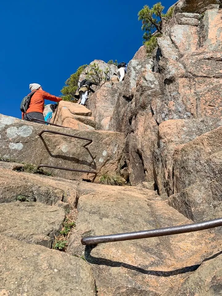 Looking up at metal bars built into rocks and lots of hikers slowly making their way up an exposed cliff