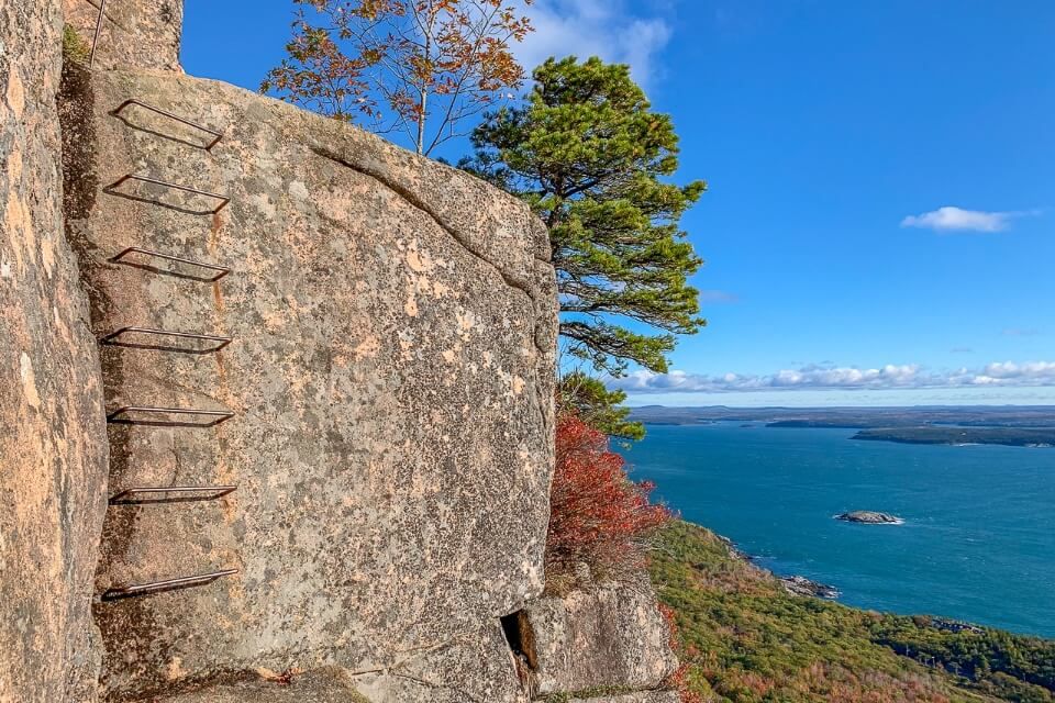 Beautiful image of an iron rung ladder in a granite rock face with views over the ocean and colorful trees precipice trail hike in acadia national park maine