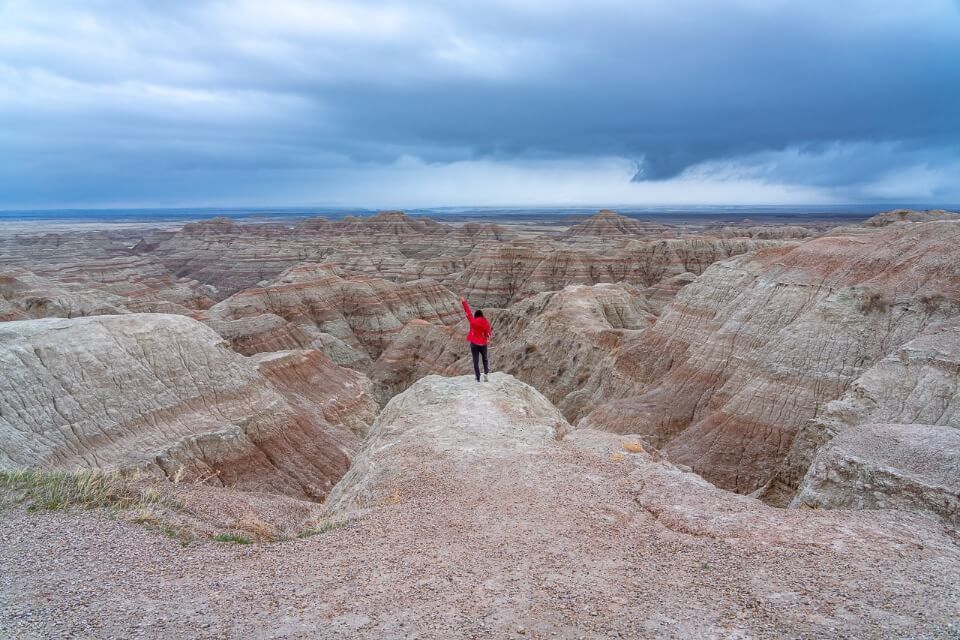Hiker raising hand in the air in front of a wide open rugged landscape with canyons and colorful soil layers beneath a dark cloudy sky