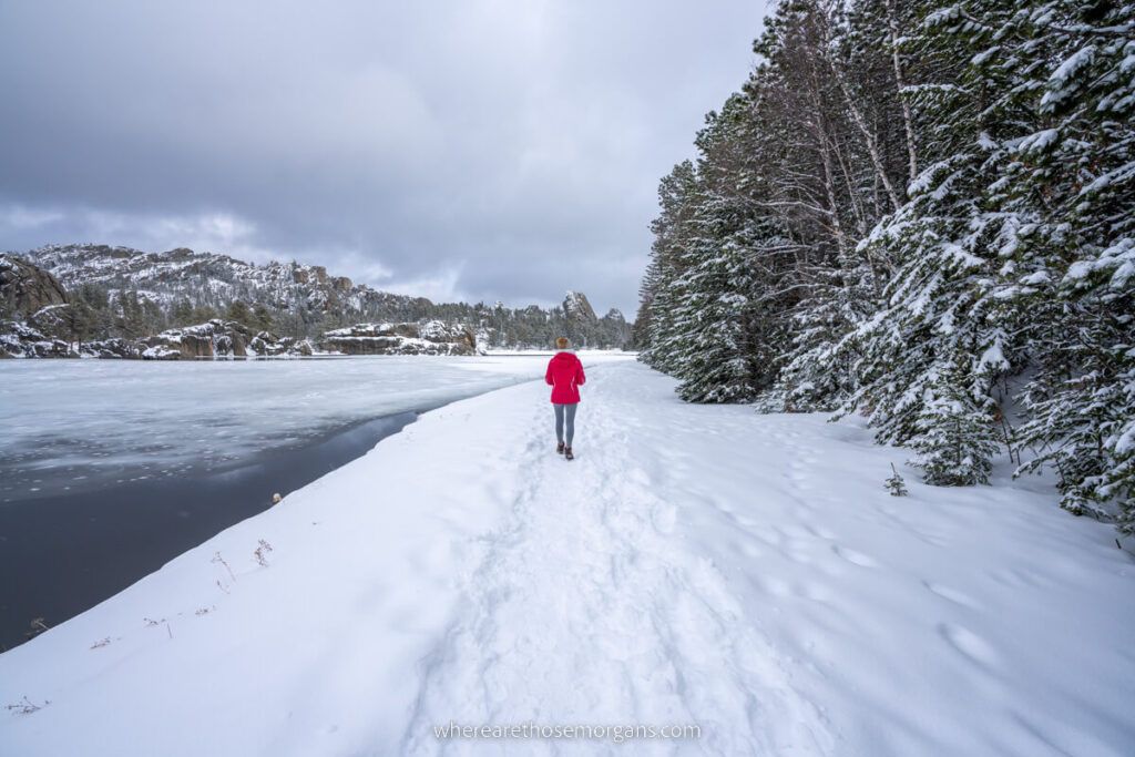 Hiker in winter coat walking on a snowy trail next to a lake and trees