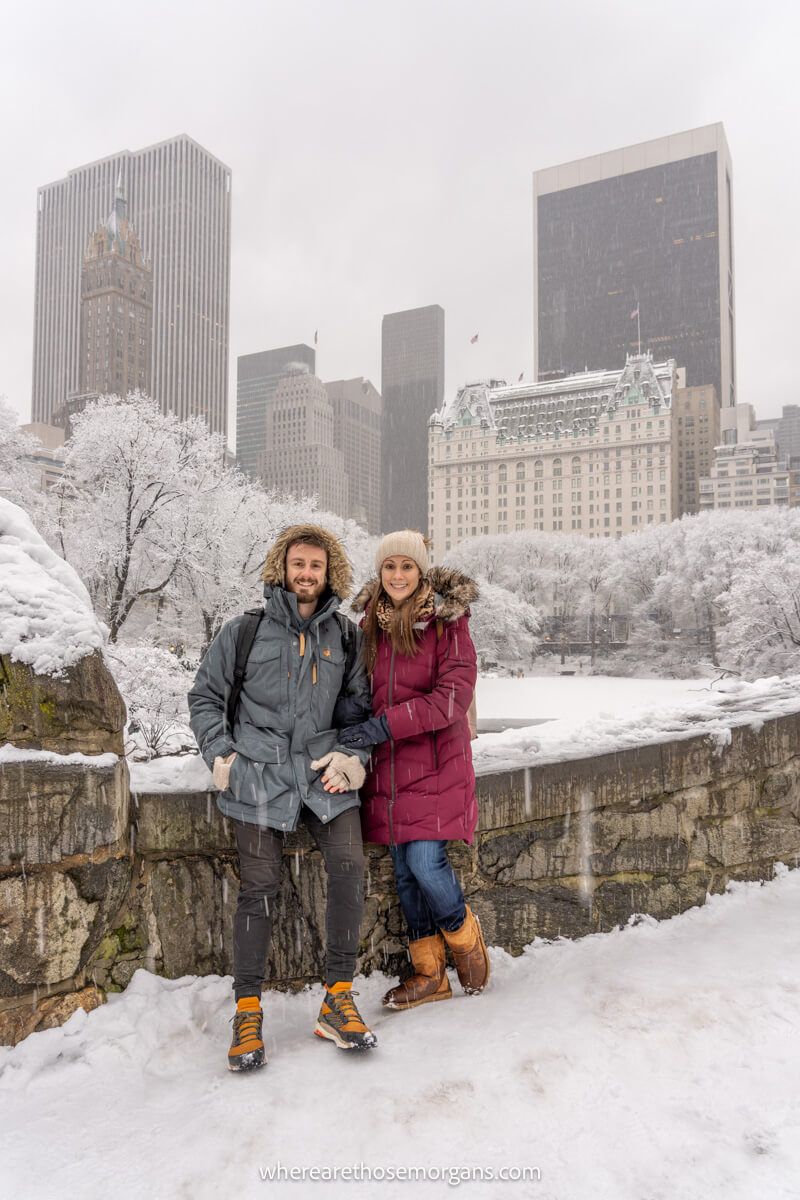 Couple in winter coats stood on a bridge in central park overlooking Fifth Avenue in heavy snow