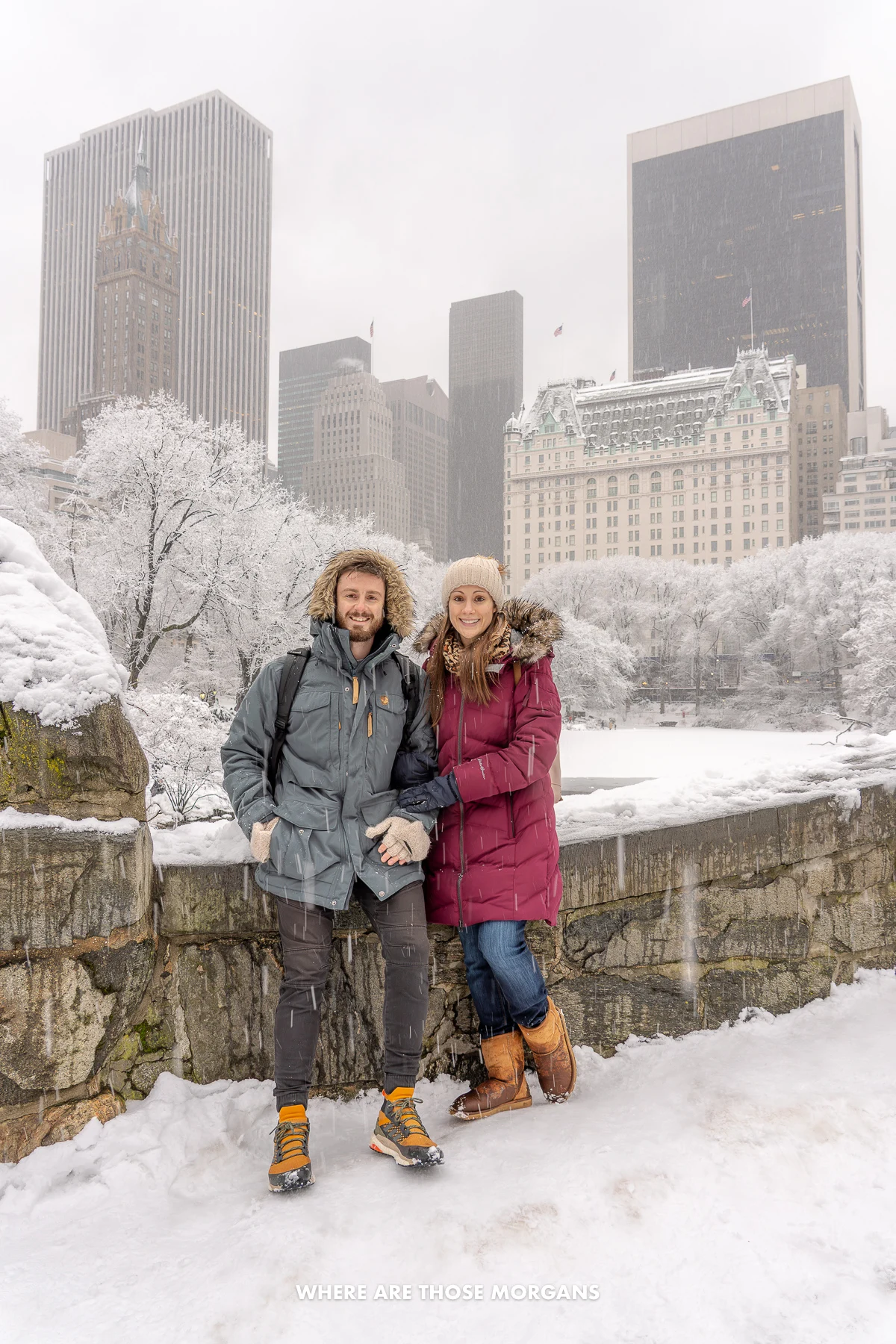 Photo of Mark and Kristen Morgan standing together in thick winter coats on a small stone bridge in Central Park covered in snow with snow falling and heavy white clouds shrouding the city's skyscrapers