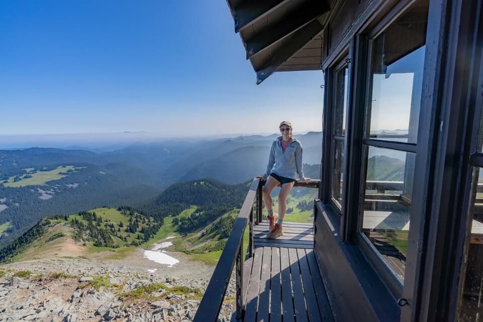 Photo of a hiker sat on a wooden rail surrounding a fire lookout tower at the top of a mountain in Mt Rainier