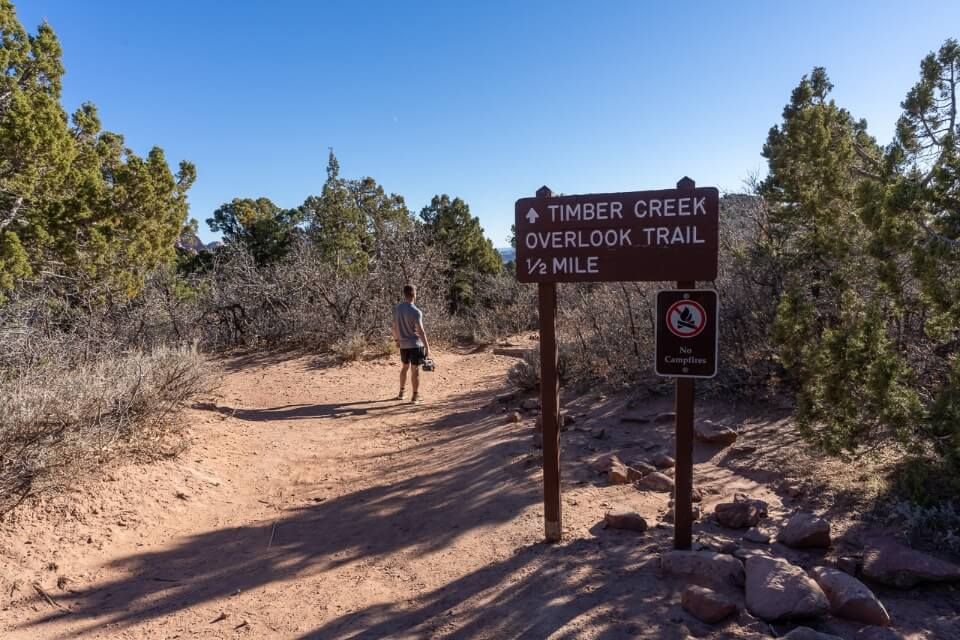 Hiker with camera equipment filming at the start of a dirt path in utah and sign stating hike distance