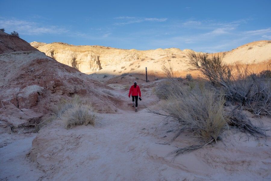 Woman hiking with red jacket in shadow on a trail leading to rocks illuminated by sunshine