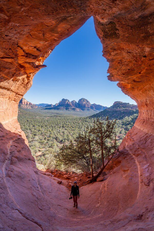 View of green trees, red rocks and blue sky with a hiker holding a camera from the inside of Birthing Cave in Sedona AZ