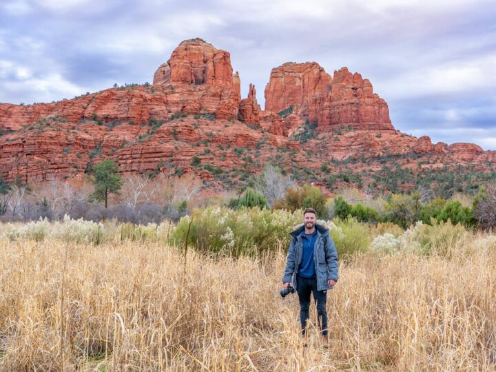Hiker with camera in field with Cathedral Rock trail in background complete guide to hiking Cathedral Rock in Sedona Arizona
