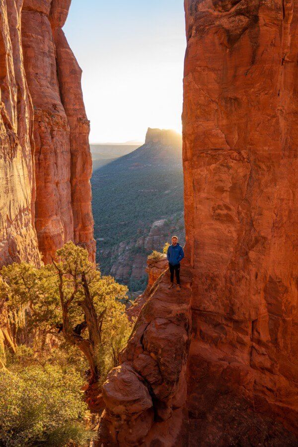 Hiker standing on distant orange rock next to a huge needle formation with the sun rising behind the rock