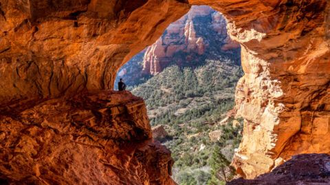 Hiker sat on ledge looking at view outside Keyhole Cave in Sedona Arizona photo taken from back of cave silhouetting hiker with bright orange glow