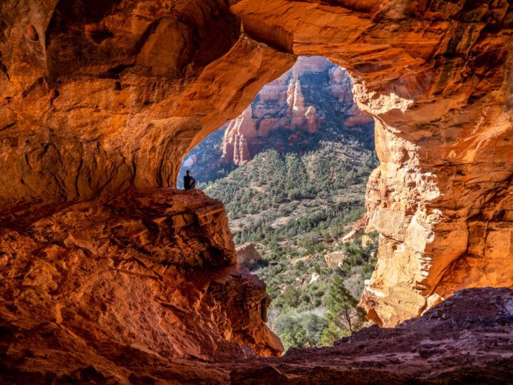 Hiker sat on ledge looking at view outside Keyhole Cave in Sedona Arizona photo taken from back of cave silhouetting hiker with bright orange glow
