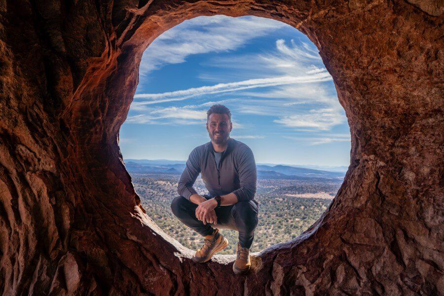 Hiker crouched in a natural sandstone window with desert landscape in background