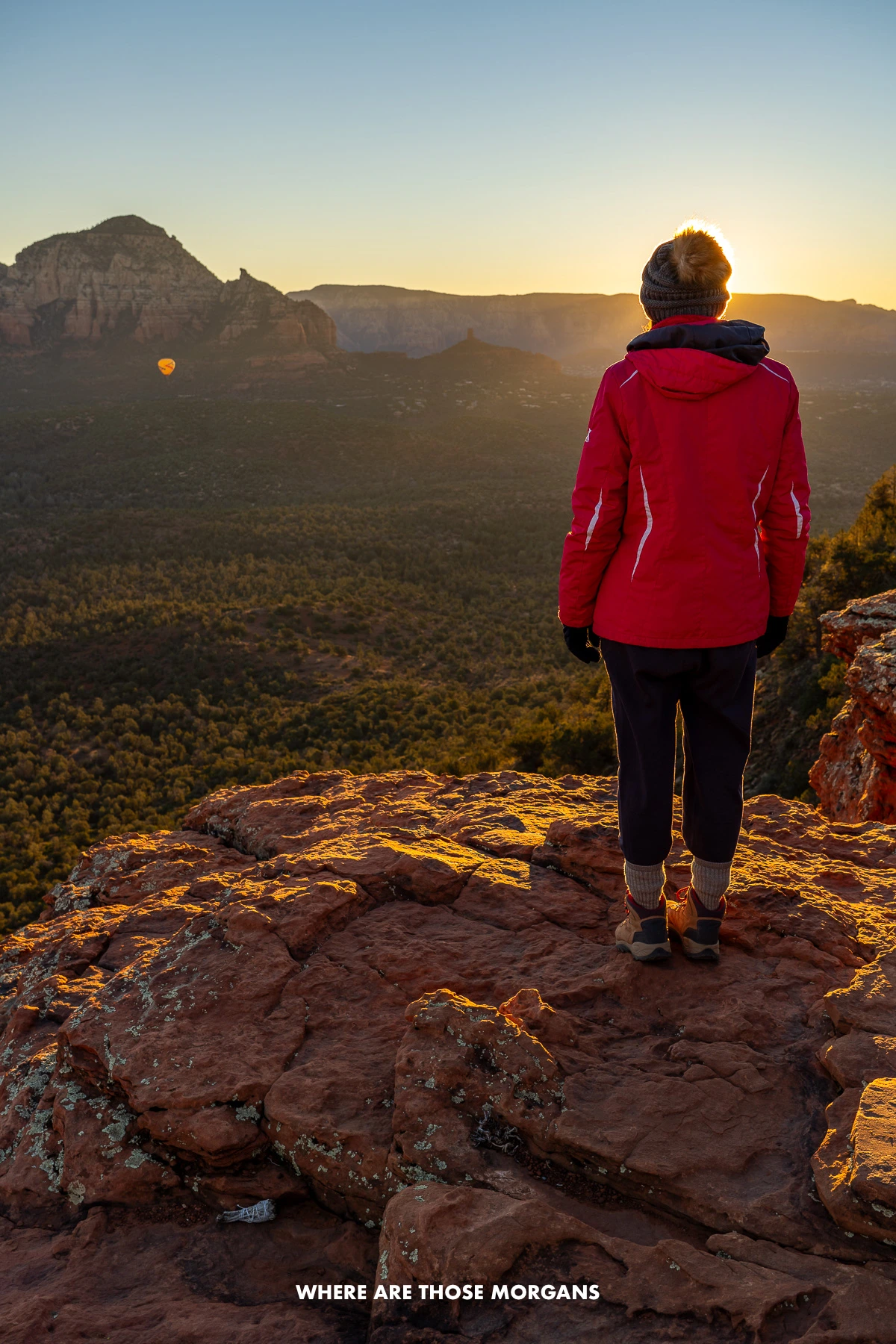 Photo of Kristen from Where Are Those Morgans standing on the edge of a red rock landscape in winter coat and hat watching a sunrise erupt over Sedona AZ from Doe Mountain summit