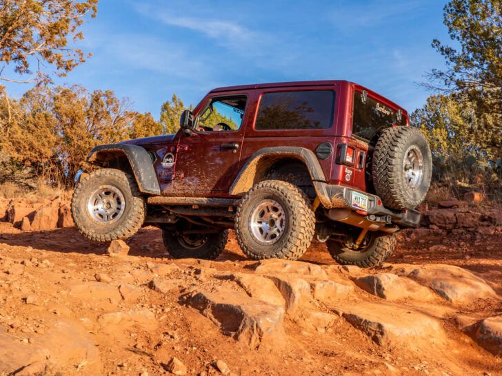 Maroon Jeep Rubicon driving over large stones on orange colored off road 4WD trail in Sedona Arizona on a sunny day in December