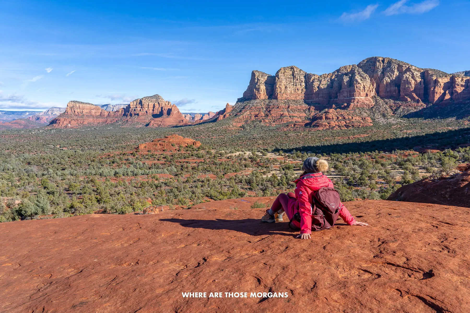 Photo of Kristen Morgan sat on a red rock shelf in a winter coat looking out at far reaching views over a valley and tall red rock cliffs under a blue sky in December