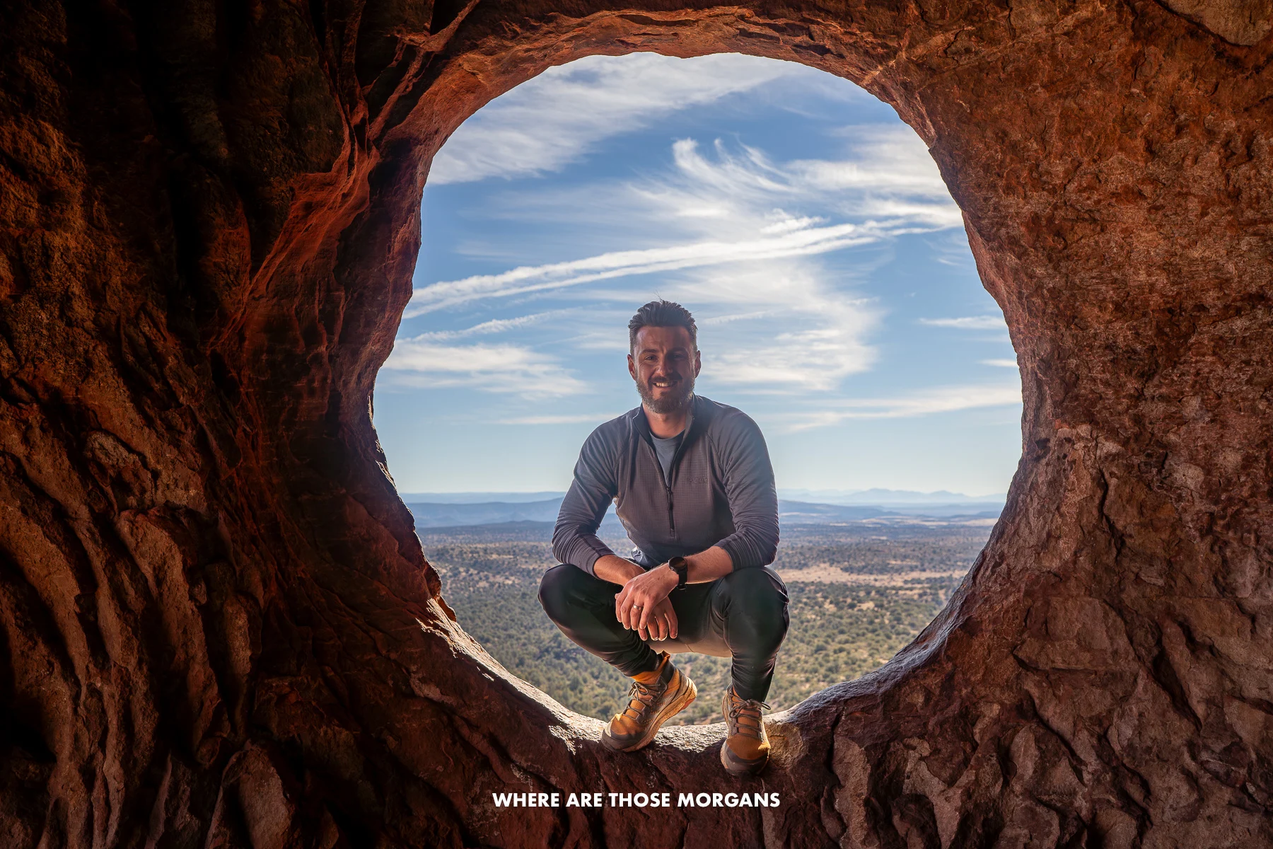 Photo of Mark Morgan crouching in a round natural window of a cave in Arizona with flat landscape views behind