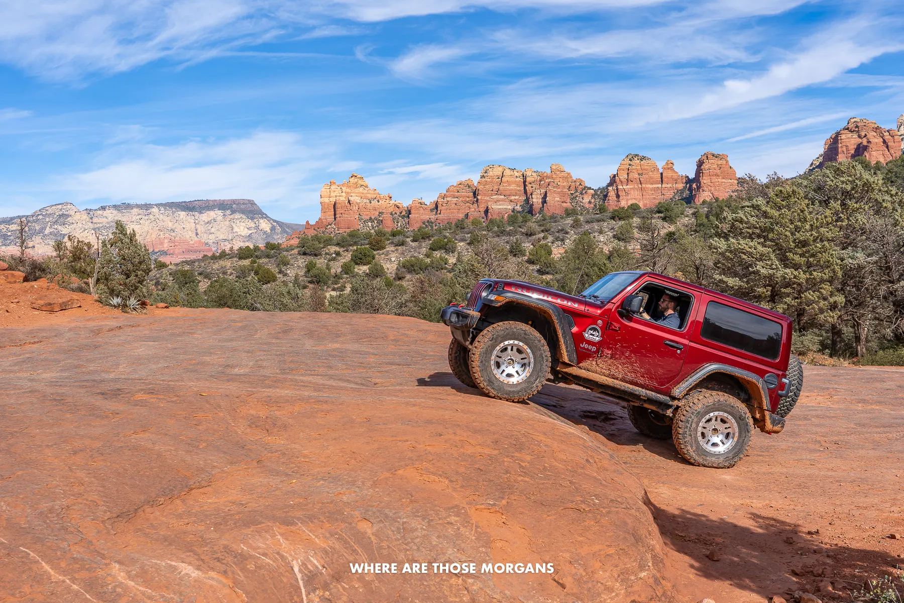 Photo of a maroon colored Jeep angled up a red rock climbing to a shelf like formation with trees and cliffs behind