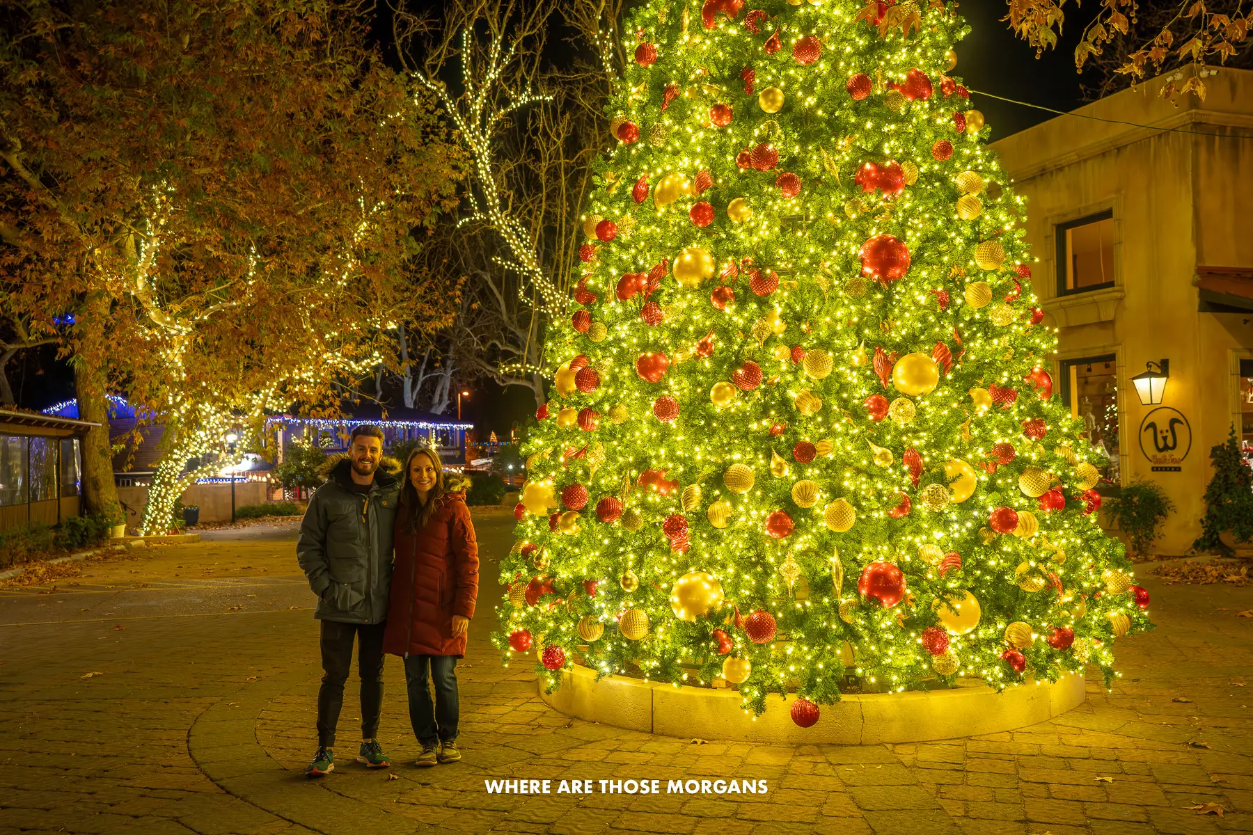 Photo of Mark and Kristen from Where Are Those Morgans stood together in winter coats next to a tall and very bright Christmas Tree in Tlaquepaque shopping village in Sedona at night in the winter