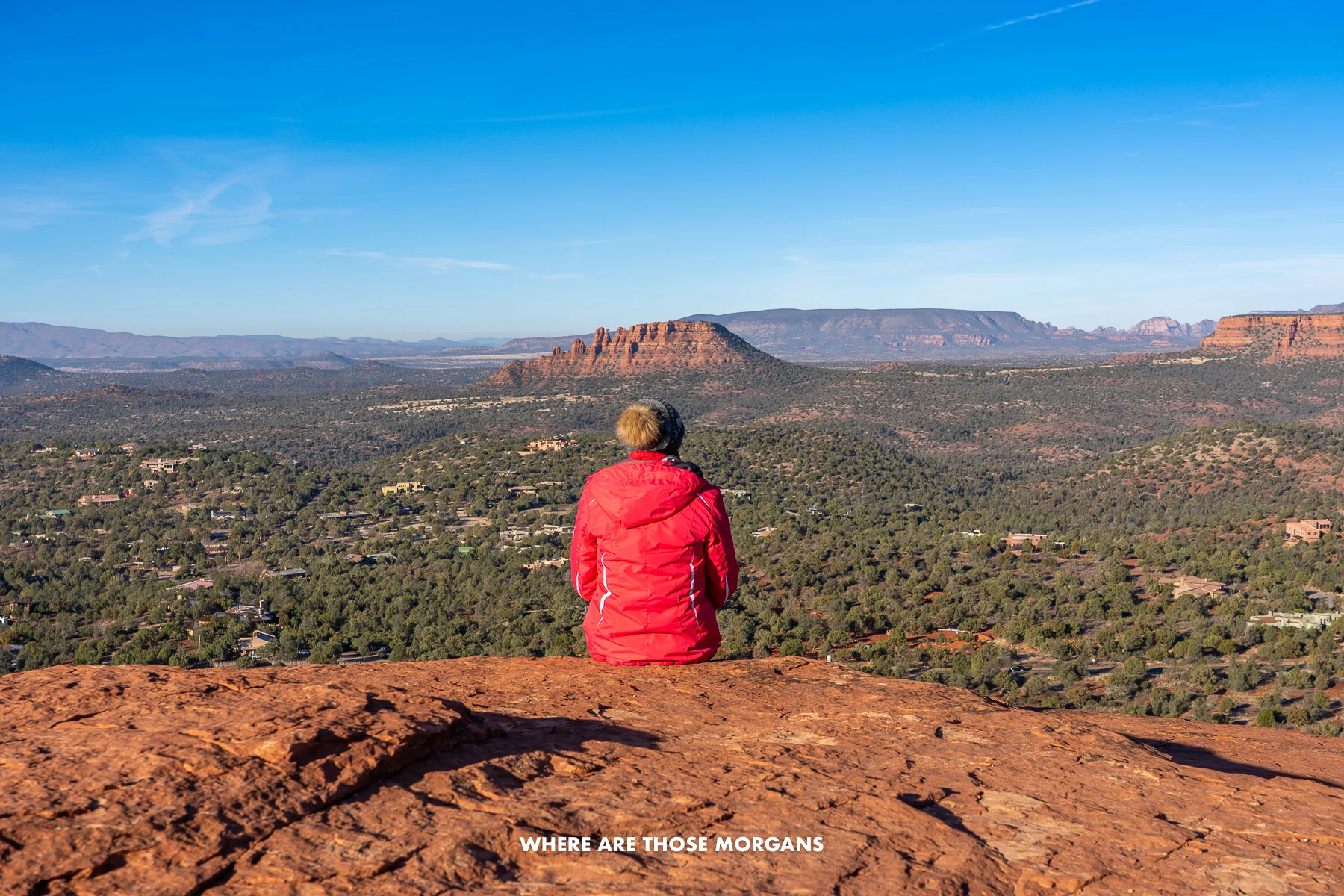Photo of Kristen Morgan sat in a coat and hat on a red rock summit looking out at far reaching views over a valley and more red rocks under a blue sky in Sedona