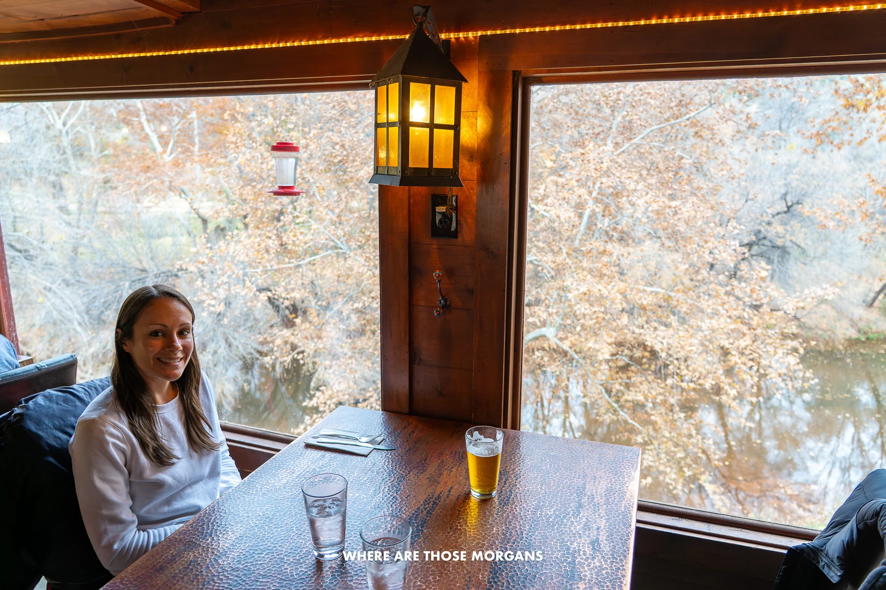 Photo of Kristen Morgan sat at a table with drinks and views of trees over water through large windows