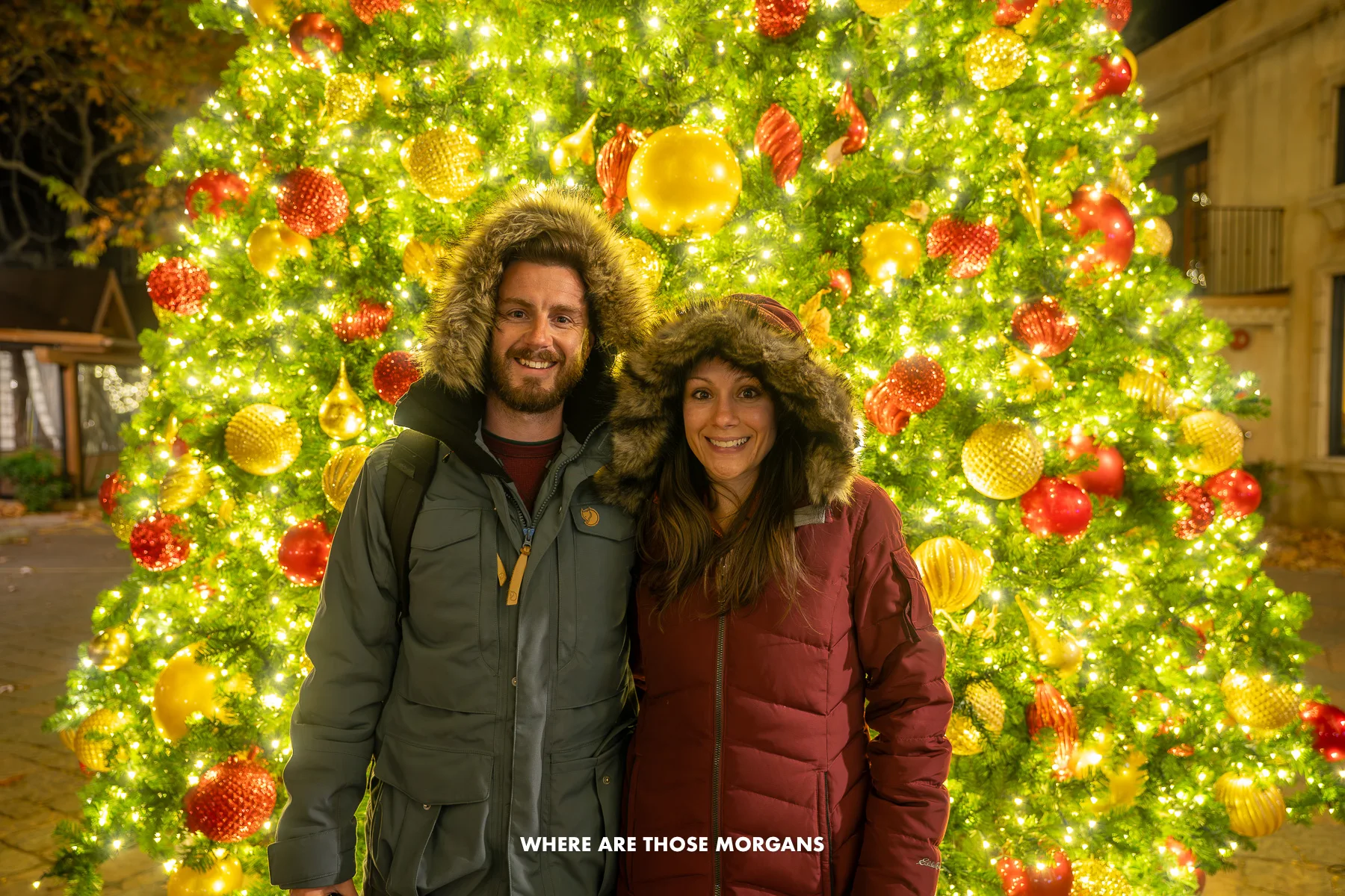 Photo of Mark and Kristen Morgan standing together in winter coats in front of a tall and wide Christmas tree in Sedona Arizona in December