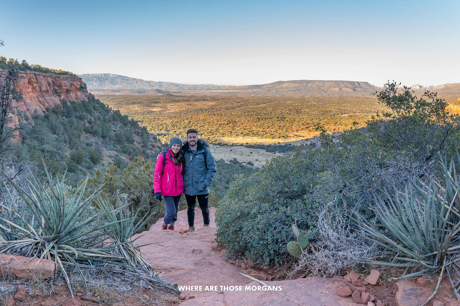 Photo of Mark and Kristen Morgan in winter coats hiking a trail in Sedona at sunrise in December with wide open landscape views behind