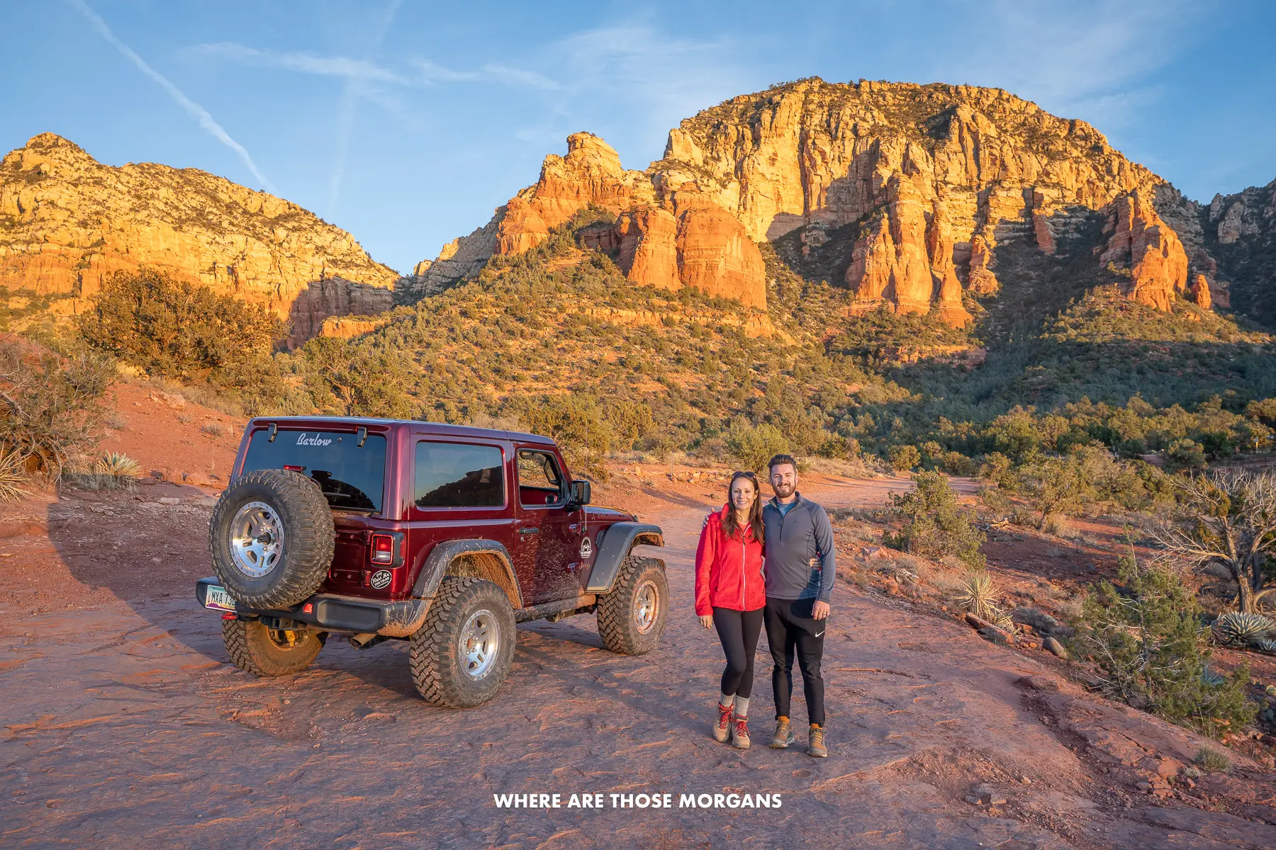 Photo of Mark and Kristen from Where Are Those Morgans standing together next to a Jeep on a red rock landscape with towering cliffs behind at sunset in Sedona