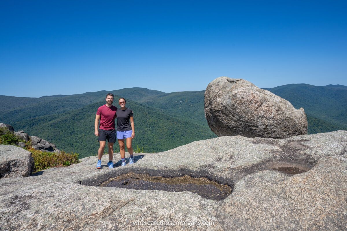 Photo of a couple standing together next to a huge boulder on a rocky surface with distant views over rolling hills on a sunny day