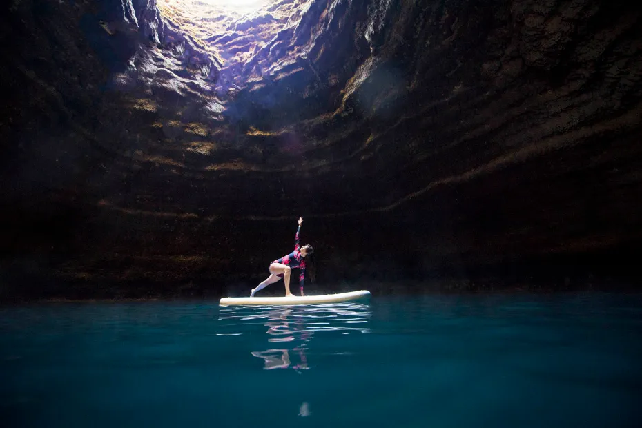 Cave with light pouring through opening above and paddle boarder on water inside cave