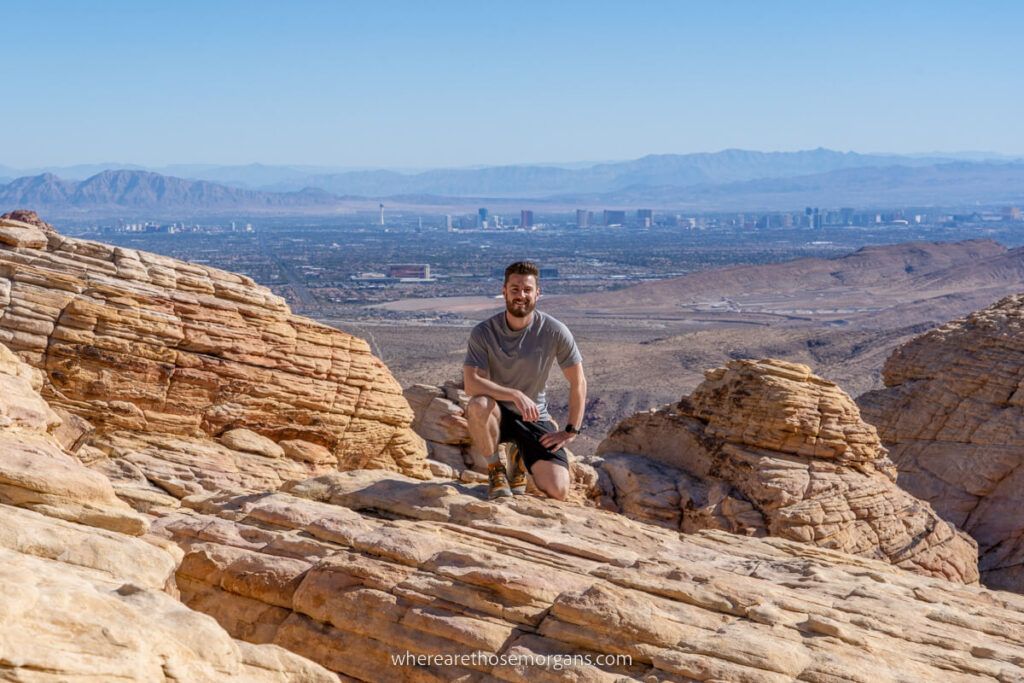 Mark from Where Are Those Morgans hiking and crouching at the summit of a trail in red Rock Canyon with Las Vegas in the distant background