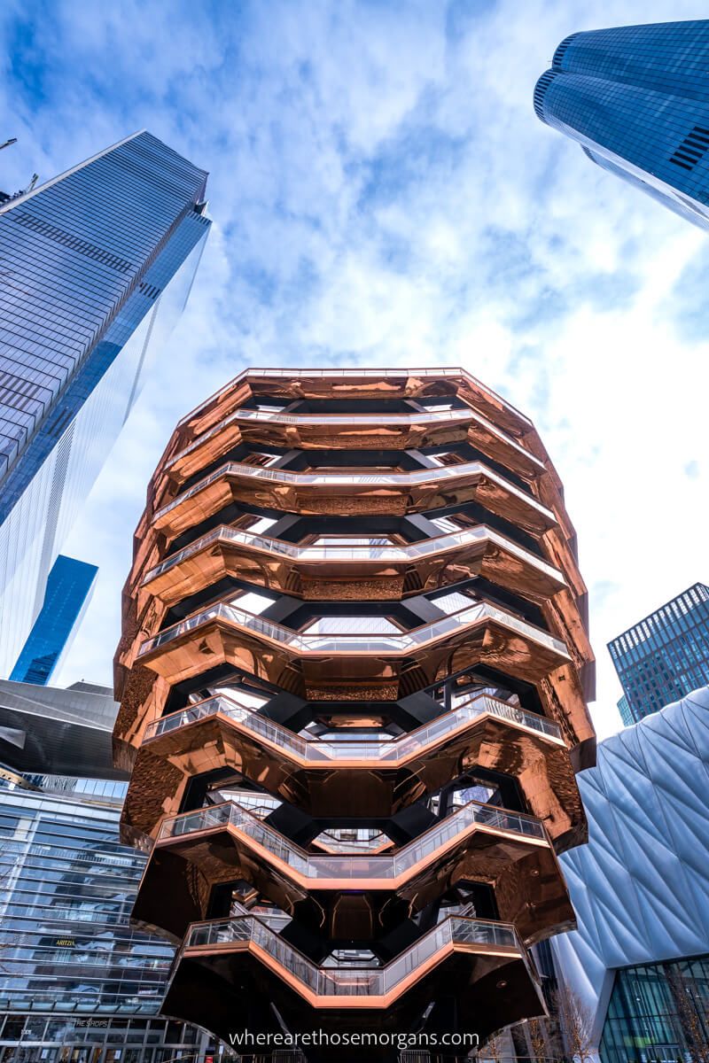 Vessel in New York City from below looking up with skyscrapers taller than the copper colored Vessel