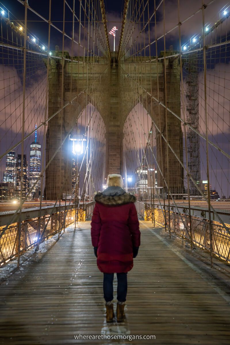 Woman walking over Brooklyn Bridge at night with maroon coat on in the center of the pedestrian walkway no crowds