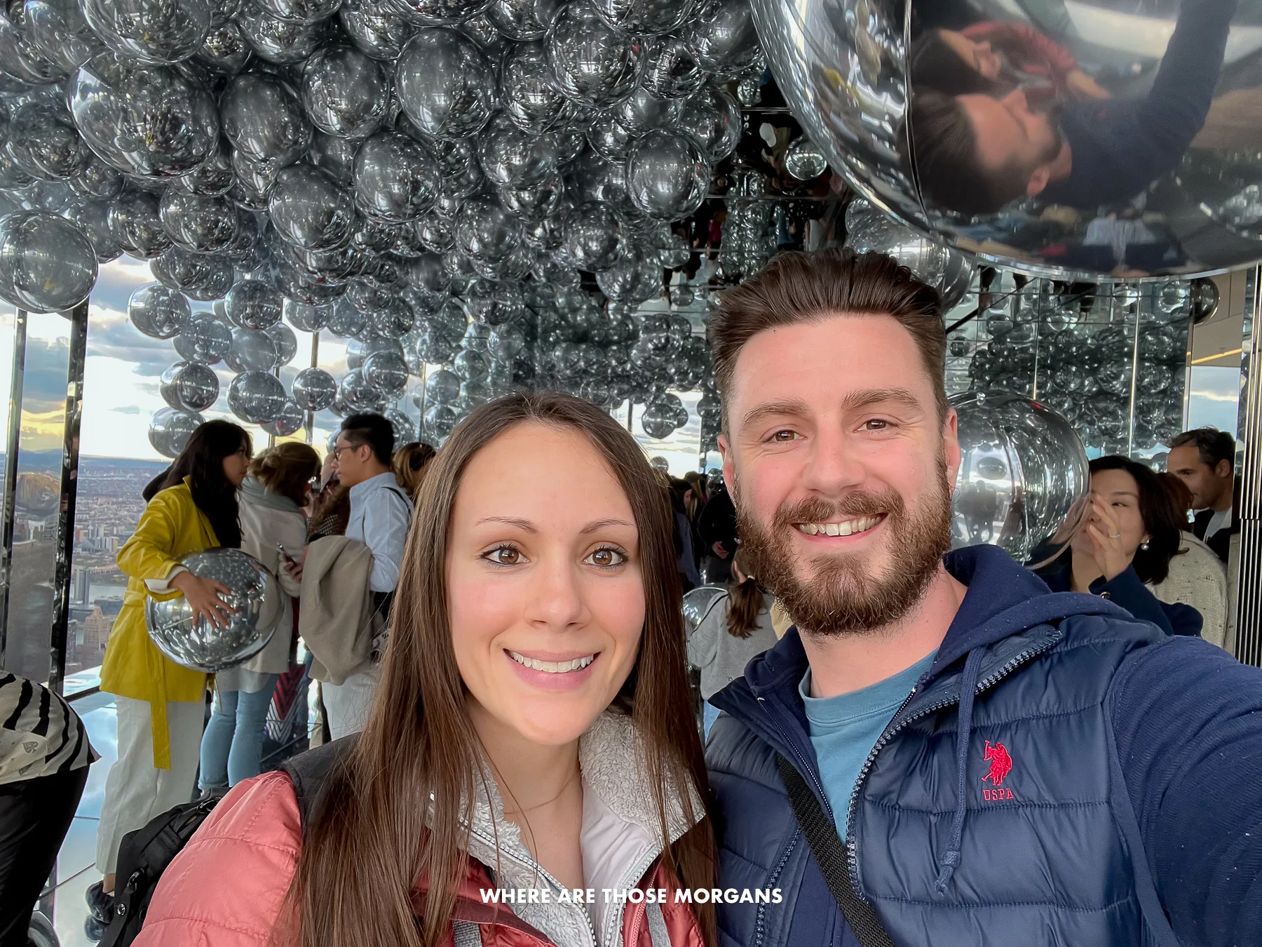 Mark and Kristen from Where Are Those Morgans taking a selfie with lots of silver helium balloons floating and tourists behind at Summit One Vanderbilt on a trip to NYC in April