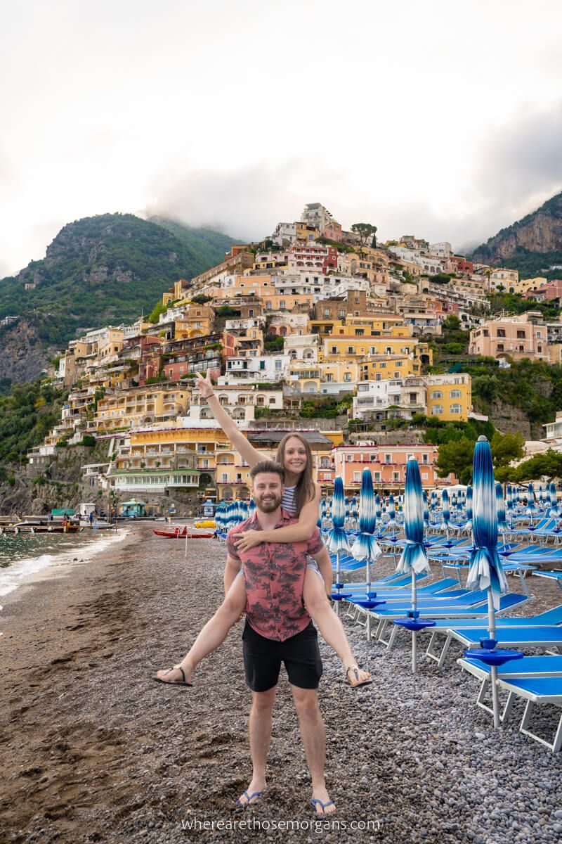Couple on Positano beach in Italy woman jumping on mans back with city on a hillside behind