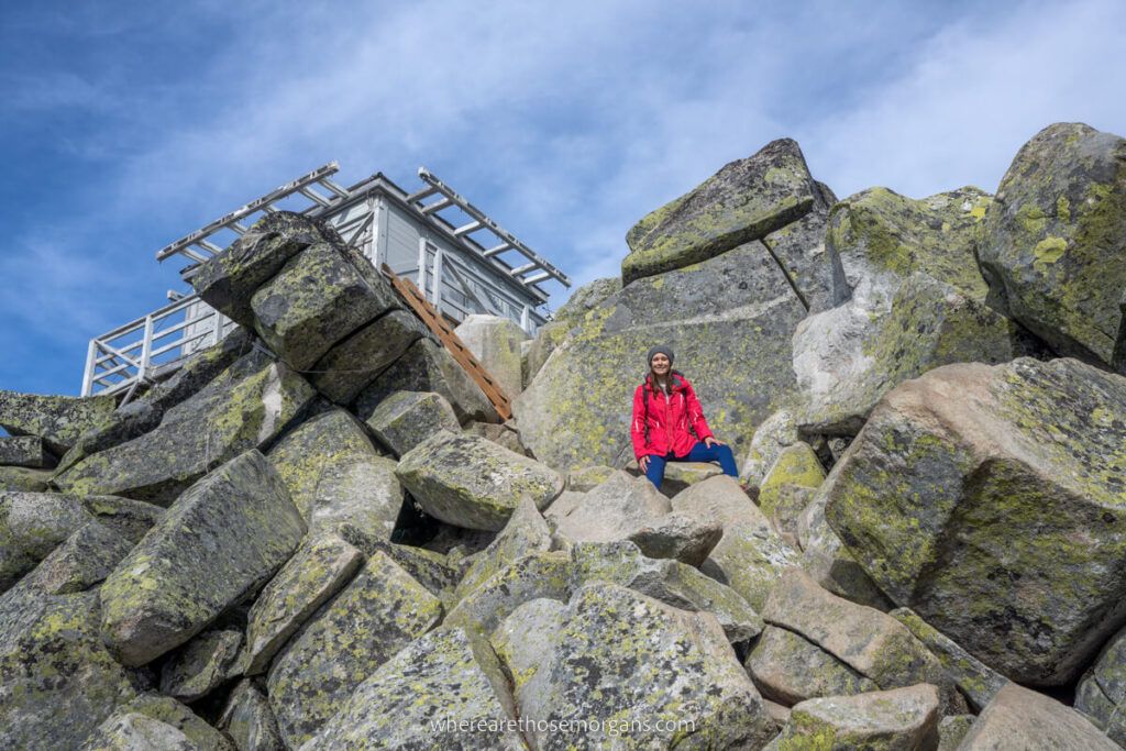 Looking up at a hiker on steep boulders with a fire lookout tower beyond