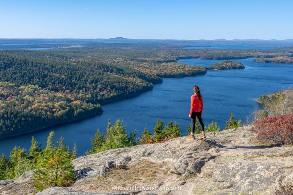 Hiker in red shirt standing on flat rocks overlooking deep blue lake and fall foliage colors in Acadia, Maine